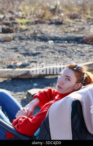 Vertikales Porträt einer rothaarigen Frau im Rollkragenpullover, liegend im Klappstuhl auf felsigem Boden, seitlich mit nachdenklichem Ausdruck. Stockfoto
