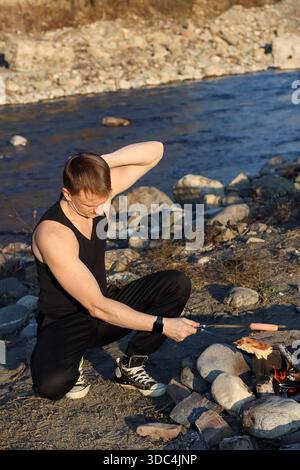 Nahaufnahme eines Mannes in sportlicher Kleidung, der sich ausdehnt, während er Wurst auf Metallspießen über glühender Lagerfeuerheuere auf Kies am Fluss kocht, was an Outdoor r erinnert Stockfoto