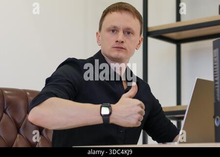 Ein junger Geschäftsmann in einem modernen Büro zeigt beim Arbeiten an einem Laptop die Daumen nach oben, was auf Zustimmung oder Erfolg hinweist. Stockfoto