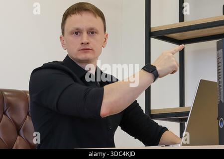 Aufmerksamer professioneller Mann am Schreibtisch, der den Zeigefinger neben den Laptop in einem modernen Büro nach oben zeigt Stockfoto