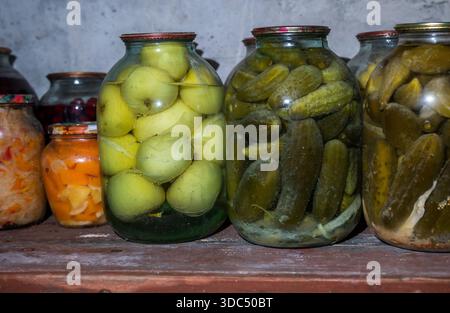 Glasgläser, die ein staubiges Holzregal auskleiden. Sorgfältige Aufbewahrung hausgemachter, fermentierter grüner Äpfel. Dill Pickles. Und verschiedene andere konservierte Früchte und Vegeta Stockfoto