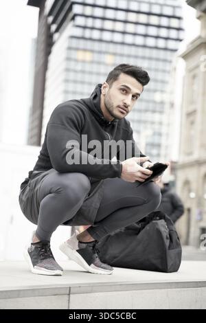 Aufmerksamer Mann, der sich mit Smartphone in der Hand in der Stadt neben der modernen Skycraper Business Tower Architektur hockt, mit nachdenklichem Geist beiseite blickt, sportlich Stockfoto