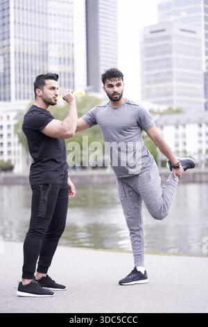 Zwei Workout-Freunde trainieren zusammen, einer macht Stretching-Übungen, während der andere Sport trinkt Elektrolyte aus der Plastikflasche, um zu bleiben Stockfoto