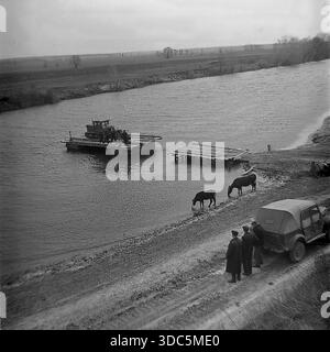 Region Donezk, ukrainische SSR, UdSSR, Mitte Ende der 1970er Jahre Stimmungsvolles Schwarz-weiß-Archivfoto einer ländlichen Flussüberquerung. Eine hölzerne Seilfähre transportiert einen schweren Raupenschlepper zur anderen Bank für Feldarbeiten. Am matschigen Ufer steht ein sowjetisches Geländefahrzeug GAZ-69, und Kühe trinken Wasser aus dem Fluss. Männer stehen in der Nähe und beobachten den Prozess. Eine Szene der landwirtschaftlichen Logistik und Infrastruktur in der abgelegenen Landschaft. Authentische Vintage-Filmfotografie des täglichen Lebens. Stockfoto