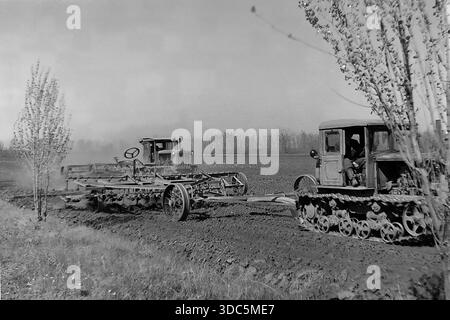 Region Donezk, ukrainische SSR, UdSSR, Mitte Ende der 1970er Jahre Atmosphärisches Schwarz-weiß-Archivfoto der Frühjahrs-Egge. Zwei sowjetische caterpillar-Traktoren (Typ DT-54 oder T-74) ziehen landwirtschaftliche Eggen über ein dunkles Pflugfeld. Junge Bäume im Vordergrund bilden die Szene und betonen die frühe Frühlingssaison. Eine klassische Betrachtung der Bodenaufbereitung und Mechanisierung in der Steppenzone. Authentische Vintage-Filmfotografie. Stockfoto