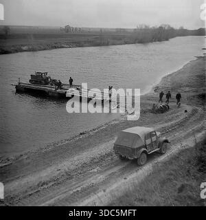 Region Donezk, ukrainische SSR, UdSSR, Mitte Ende der 1970er Jahre Stimmungsvolles Schwarz-weiß-Archivfoto einer ländlichen Flussüberquerung. Eine hölzerne Seilfähre transportiert einen schweren Raupenschlepper zur anderen Bank für Feldarbeiten. Am matschigen Ufer steht ein sowjetisches Geländefahrzeug GAZ-69, und Kühe trinken Wasser aus dem Fluss. Männer stehen in der Nähe und beobachten den Prozess. Eine Szene der landwirtschaftlichen Logistik und Infrastruktur in der abgelegenen Landschaft. Authentische Vintage-Filmfotografie des täglichen Lebens. Stockfoto