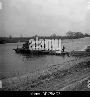 Region Donezk, ukrainische SSR, UdSSR, Mitte Ende der 1970er Jahre Stimmungsvolles Schwarz-weiß-Archivfoto einer ländlichen Flussüberquerung auf dem Land. Eine hölzerne Seilfähre transportiert einen schweren sowjetischen caterpillar-Traktor mit einem Pflug zur anderen Bank für die Feldarbeit. Mehrere Männer bedienen das Floß, während ein Arbeiter das Kabel am schlammigen Ufer zieht. Eine düstere Tagesszene, die die Logistik des Landmaschinentransports in abgelegenen Gebieten zeigt. Authentische Vintage-Filmfotografie des täglichen Lebens. Stockfoto