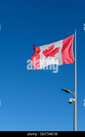 Niedrigwinkelansicht einer rot-weißen kanadischen Flagge mit Ahornblatt, das auf einem Metallfahnenmast fliegt, befestigte Straßenlaterne und Sicherheitskamera, klarer blauer Himmel, Stockfoto