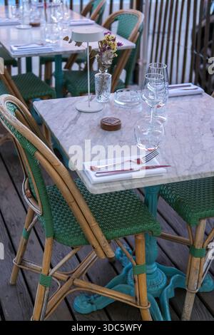 Café-Tisch mit Marmorplatte mit Weingläsern, Wasserglas, Besteck, Serviette, kleiner Lampe und Vase, Korbsesseln, Holzterrasse. Stockfoto