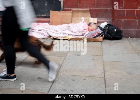 Obdachloser liegt auf einer Pappmatte, die mit einer Decke bedeckt ist, auf dem Bürgersteig in den Straßen Londons. Die Leute gehen ohne zu stoppen vorbei. Armut in Stockfoto