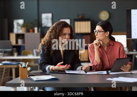Zwei junge Erwachsene Frauen sitzen am Schreibtisch und diskutieren Dokumente und ein digitales Tablet in einem modernen Büroarbeitsplatz Stockfoto