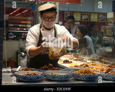 Peking-Peking-China-14. August 2018, ein asiatischer Lebensmittelhändler, trägt eine Maske und Schürze mit der Aufschrift „老街佬“, bereitet vorgekochte traditionelle chinesische Gerichte (Lu Stockfoto