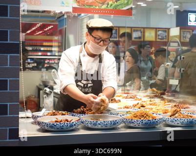 Peking-Peking-China-14. August 2018, ein asiatischer Lebensmittelhändler, trägt eine Maske und Schürze mit der Aufschrift „老街佬“, bereitet vorgekochte traditionelle chinesische Gerichte (Lu Stockfoto