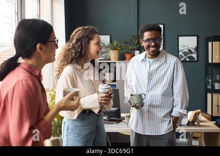 Multiethnische Gruppe junger Erwachsener, die zusammen Kaffeetassen halten, lächeln und reden in einem modernen Büroumfeld, Schwarzer Mann und zwei kaukasische Frauen, die sich unterhalten Stockfoto
