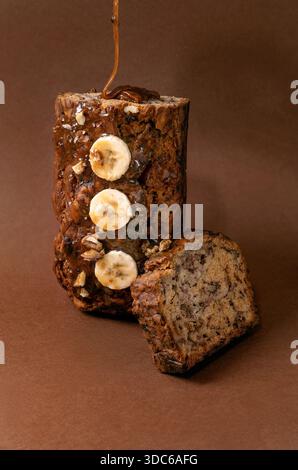 Modernes Stillleben mit hausgemachtem Bananenbrot. Brot mit Walnüssen, mit Karamell getränkt, aufrecht stehend auf braunem Hintergrund. Soße von oben ausgießen Stockfoto