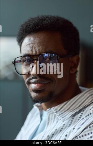 Porträt des Schwarzen Mannes mit Brille, der mit einem leichten Lächeln in die Kamera blickt, kurze lockige Haare und beschnittener Spitzbart sichtbar, Inneneinrichtung mit verschwommenem Hintergrund Stockfoto