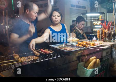 hanoi-Vietnam-10. August 2019, Hanoi, Vietnam. Straßenhändler bereiten gegrillte Spieße und Bánh Mì-Würstchen an einem von Neonlichtern beleuchteten Stand zu Stockfoto