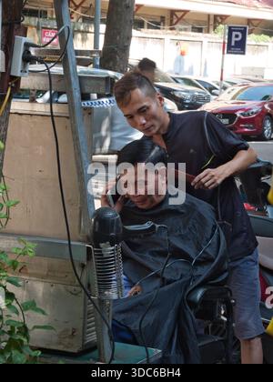 hanoi-Vietnam-9. August 2019, ein informeller Straßenfriseur schneidet einem älteren Mann die Haare, der mit seinem Telefon spricht, und hebt das tägliche Leben und das traditionelle t hervor Stockfoto
