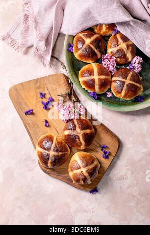 Heiße Kreuzbrötchen auf Holzbrett und Keramikplatte. Traditionelles Ostergebäck mit Frühlingsblumen. Hausgemachtes Süßbrot mit Hochglanzkruste Stockfoto