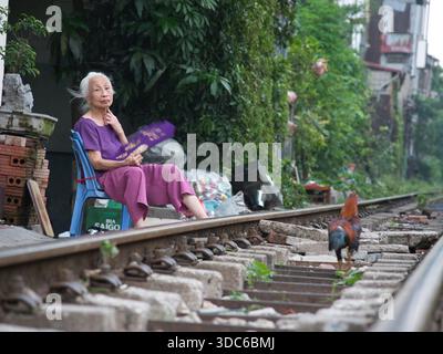 hanoi-Vietnam-10. August 2019, eine ältere vietnamesische Frau sitzt auf einem Plastikstuhl neben aktiven Bahngleisen und fächert sich in einer städtischen Residentia an Stockfoto