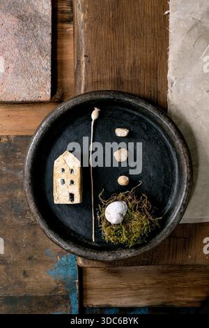 Nahaufnahme schwarze Keramikschale mit Miniatur-Keramikhaus, kleine Kieselsteine, Moosnester mit Schale auf rustikalem Holzhintergrund. Naturmaterialien künstlerisch Stockfoto