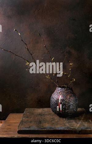 Minimales Stillleben mit aufblühenden Frühlingszweigen in kunstvoller Keramikvase. Dunkelbrauner, strukturierter Hintergrund. Rustikaler Holztisch. Stimmungsvolle Atmosphäre. Stockfoto