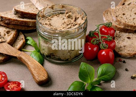 Hausgemachte Leberpastete im Glasgefäß mit Brotscheiben Kirschtomaten, frischen Basilikumblättern und Holzlöffel. Gesunde Vorspeise zum Frühstück oder Snack. Stockfoto