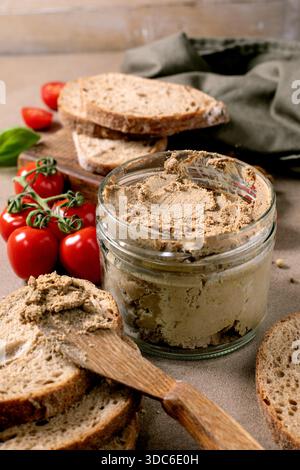 Hausgemachte Fleischpastete im Glas, serviert mit frischen Vollkornbrotscheiben, Kirschtomaten und Basilikumblättern. Rustikaler Holzlöffel und Schneidebrett. Stockfoto