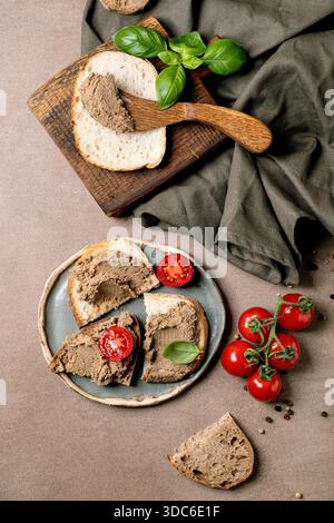 Hausgemachte Leberpastete auf Brotscheiben serviert mit frischen Kirschtomaten Basilikumblättern auf Holzbrett und Keramikplatte. Vorspeise im rustikalen Stil. Stockfoto