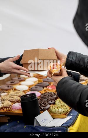 Person, die bunte Donuts und Gebäck am Street Food Stand kauft. Hände teilen sich Bastelbox mit süßen Leckereien. Verschiedene glasierte Donuts auf dem Präsentationsfach. C Stockfoto