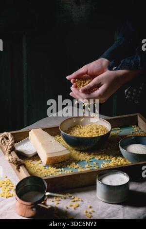 Junger Mann in Schwarz, der rohe ungekochte italienische Pasta-Coquillettes zum Kochen des Abendessens mit Parmesankäse, Sahne und Grießmehl auf Holztablett auf Küchen-Leinen-Tischtuch bestreut. Dunkler, rustikaler Stil. Stockfoto