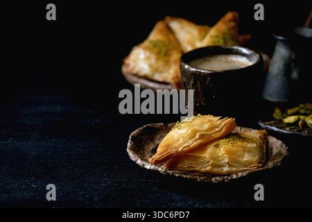 Hausgemachtes türkisches traditionelles Dessert Baklava mit Pistazien serviert auf Keramikplatte mit Nüssen, Tasse Kaffee, Vintage-Cezva auf schwarzem Holztisch. Kopierbereich Stockfoto