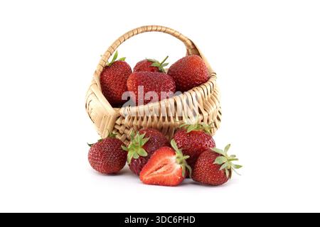 Reife rote Erdbeeren fließen aus einem kleinen Korb auf einen sauberen weißen Hintergrund und schaffen ein lebendiges und appetitliches Schauspiel mit einer Hälfte Erdbeere, die das saftige Interieur unterstreicht Stockfoto