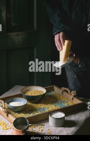 Junger Mann in schwarzem Käse. Rohe, ungekochte italienische Pasta Coquillettes zum Kochen des Abendessens mit Parmesan, Sahne und Grießmehl auf Holztablett auf Küchenwäsche Tischtuch. Dunkler, rustikaler Stil. Stockfoto