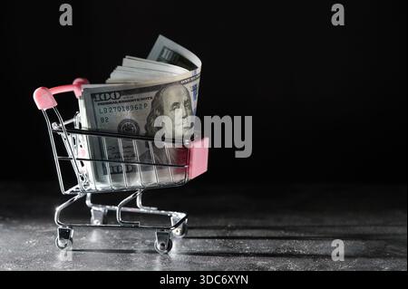 Warenkorb auf Rädern mit Dollarscheine auf dunklem Hintergrund, Nahaufnahme, Kopierraum. Finanz-, Geschäfts-, Einkaufs-, Ausgaben- und Vertriebskonzept Stockfoto