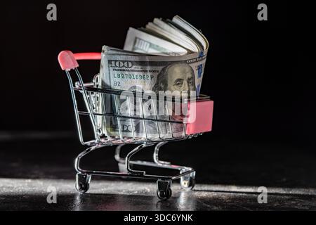 Warenkorb auf Rädern mit Dollarscheine auf dunklem Hintergrund, Nahaufnahme, Kopierraum. Finanz-, Geschäfts-, Einkaufs-, Ausgaben- und Vertriebskonzept Stockfoto