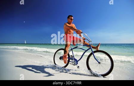 Ein Mann fährt mit dem blauen Fahrrad an einem Sandstrand. Er trägt rote Shorts und tritt durch seichtes Wasser. Der Himmel ist klar und sonnig. Wellen krachen sanft in t Stockfoto