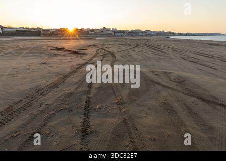 Ein ruhiger Strand bei Sonnenuntergang zeigt Reifenspuren, die den Sand in Richtung einer entfernten Küstenstadt Buzowna überqueren. Das warme Leuchten der Sonne erzeugt lange Schatten, Invi Stockfoto