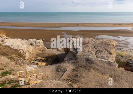Eine ruhige Küstenlandschaft mit Holztreppen, großen Felsen, einer Sandküste und einem ruhigen Blick auf das Kaspische Meer unter weichem Himmel Stockfoto
