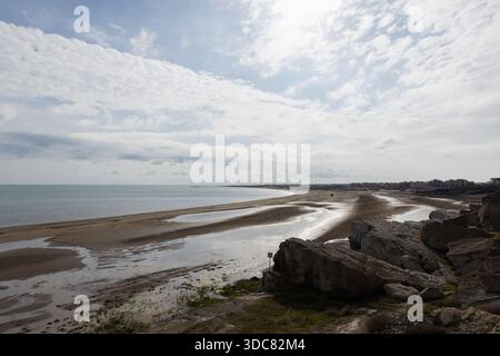 Eine ruhige Küstenlandschaft mit einem langen Sandstrand, ruhigem Wasser, felsigem Vorderboden und einer entfernten Küstenstadt. Weiche Wolken driften darüber und erzeugen Stockfoto