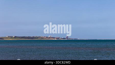 Ein ruhiges Panorama an der Küste zeigt ein breites, blaues Meer, das auf eine ferne Küste mit Gebäuden trifft. Klarer Himmel und ruhige Atmosphäre erinnern an Ruhe Stockfoto
