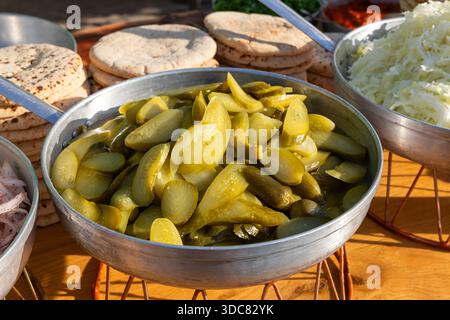 Metallschale gefüllt mit geschnittenen eingelegten Gurken in Salzlake auf einer Holztheke neben gehacktem Kohl und Pita-Stacks in einem zwanglosen Café, bereit als würzige Burger-Toppings während des Mittagessens Stockfoto