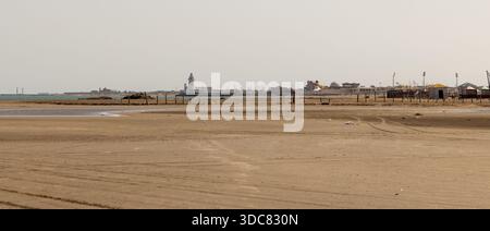 Ruhige Küstenszene mit ausgedehntem Sandstrand, ruhigem Wasser, einem entfernten Pier und einem Leuchtturm unter klarem Himmel. Küste des Kaspischen Meeres, Buzowna, Stockfoto