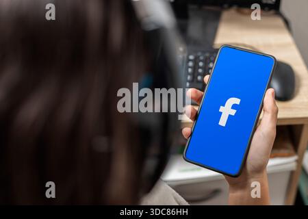 Ein nicht erkennbarer weiblicher Teenager hält ein Mobiltelefon mit einem Facebook-Logo auf dem Bildschirm mit blauem Hintergrund Stockfoto