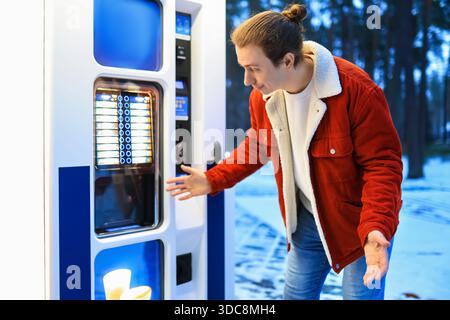 Verwirrter junger Mann in roter Jacke, der am Kaffeeautomaten im verschneiten Winterpark Gesten macht. Hochwertige Fotos Stockfoto