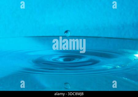 Minimalistischer Hochgeschwindigkeitsabfang eines einzelnen Wassertropfens, der über einer ruhigen Oberfläche aufliegt, erzeugt glatte, konzentrische Wellen in einer blau gefärbten Umgebung Stockfoto
