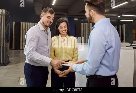 Junger, fröhlicher Mann, der Kunden, die auf dem Baumarkt stehen, Fliesen zeigt. Stockfoto