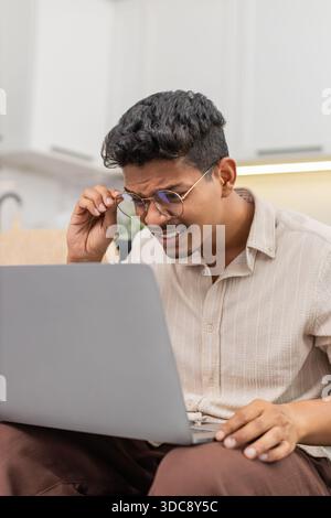 Ein indischer Mann zu Hause hat Schwierigkeiten, winzige Texte auf dem Laptop zu lesen, zieht dann eine Brille auf und sieht klare Sicht. Arabian Guy on Sofa Lächeln passt den Rahmen an, liest Bildschirmlinien ohne Belastung und setzt die Arbeit fort. Stockfoto