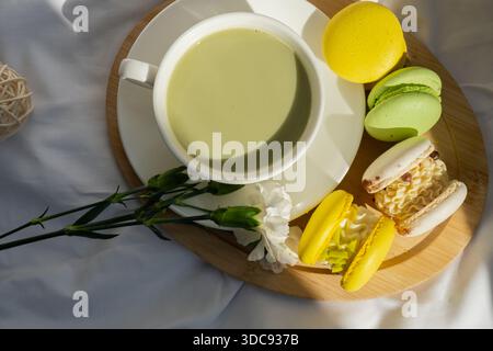 Süßes gelbes Macaron Dessert Tasse grüner Matcha Tee Latte auf Pflanzenbasis in gemütlicher Morgenliegenatmosphäre. Kreative Urlaubsidee mit romantischem Date. Pflanzliche präbiotische Faser Superfood Getränk Antioxidans-reiche Immunität-Boosting Kaffee Alternativen Getränk Stockfoto