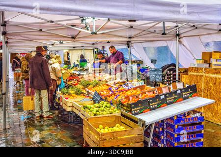 Käuferinnen und Käufer wählen Gemüse aus einem überdachten Verkaufsstand auf dem Freiluftmarkt an regnerischen Tagen - Loches, Indre-et-Loire (37), Frankreich. Stockfoto